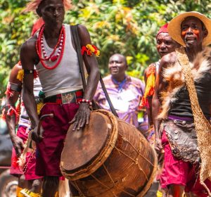 Group of African men in traditional attire performing with drums during a vibrant outdoor cultural festival.